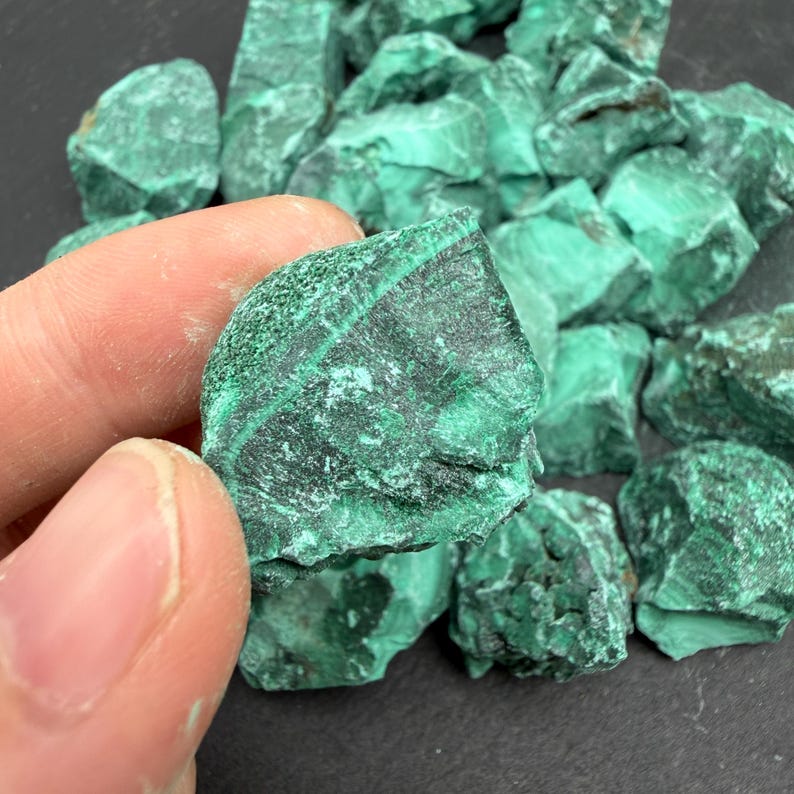 Op de afbeelding: Een verzameling ruwe malachietstenen met diep bosgroene en lichtere smaragdgroene banden. De stenen hebben een ruwe, kristallijne textuur met zichtbare lagen en natuurlijke mineraalpatronen over de grillige, onregelmatige oppervlakken.