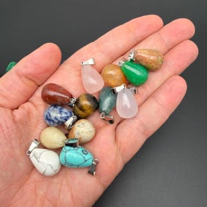 May include: A collection of teardrop-shaped gemstone pendants in various colors including white, turquoise, blue, brown, yellow, green, and pink. Each polished stone features a silver-toned metal bail attached to the top for hanging as a necklace.