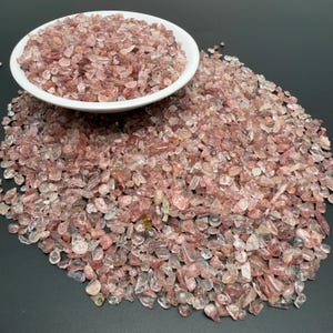 May include: A white ceramic bowl filled with small, tumbled strawberry quartz gemstone chips. A large pile of the same translucent, pinkish-red mineral fragments is spread out around the base of the bowl, showcasing varied shapes and sizes.