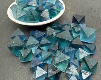 May include: A collection of translucent blue and teal fluorite crystals shaped as octahedrons. Some crystals are contained within a small white bowl, while others are scattered across the surface. The stones exhibit varying shades of blue.