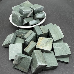 May include: A collection of sage green stone cubes with natural variations in texture and colour. Some cubes feature thin white or brown mineral veins. A portion of the stones are piled inside a white bowl, while others are scattered around the base.