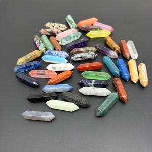 Pode incluir: Uma coleção de pontas de cristal de terminação dupla em várias cores e texturas, incluindo pedras sólidas, translúcidas e manchadas. Estes pequenos espécimes minerais alongados de forma hexagonal estão espalhados sobre uma superfície lisa.