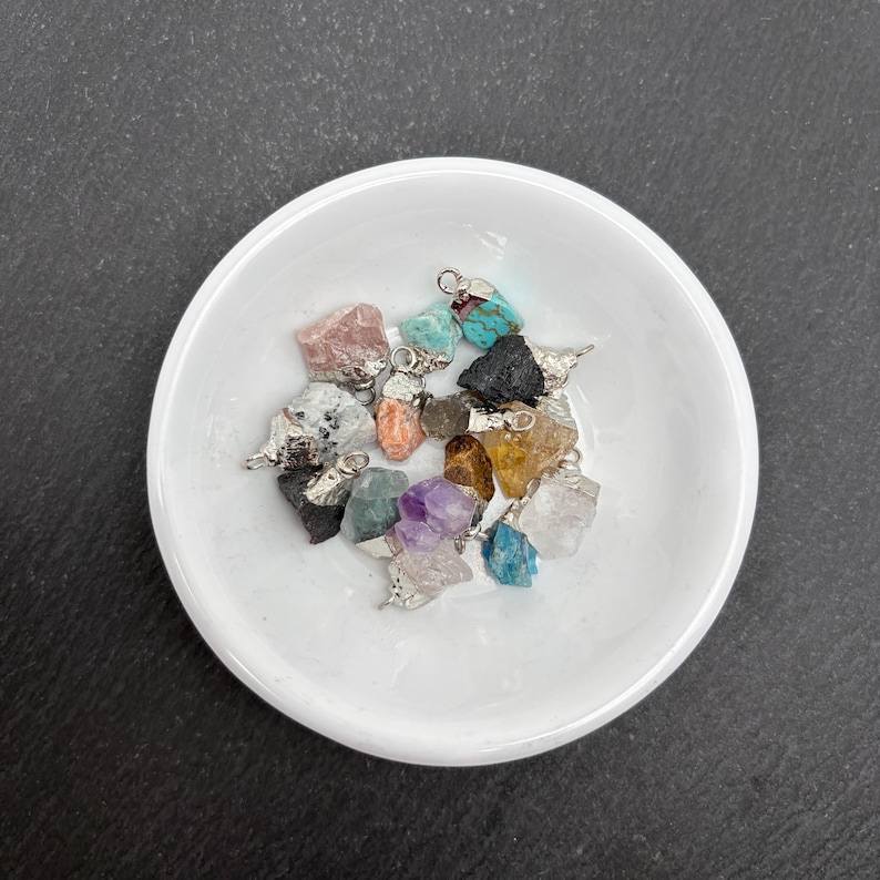 Pu&ograve; includere: Una collezione di piccoli ciondoli in pietre preziose grezze di vari colori, tra cui turchese, ametista, quarzo rosa e quarzo trasparente. Ogni pietra grezza &egrave; dotata di un gancio in metallo argentato sulla parte superiore come ciondolo.