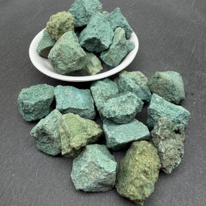 May include: A collection of raw, unpolished green stones with rough, jagged textures and varying shades of sage, mint, and forest green. Some stones are contained within a white ceramic bowl, while others are scattered around the base.