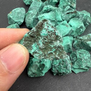 Op de afbeelding: Een verzameling ruwe malachietstenen met levendige groene tinten en donkere, getextureerde mineraalpatronen. De stenen hebben een onregelmatige, grillige vorm met een ruw, kristallijn oppervlak dat natuurlijke variaties in kleur en diepte vertoont.