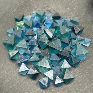 May include: A collection of numerous small, translucent blue and teal fluorite crystals, each shaped as an octahedron with sharp geometric edges and smooth, glassy surfaces, clustered together to display varying shades of deep ocean blue and light green.