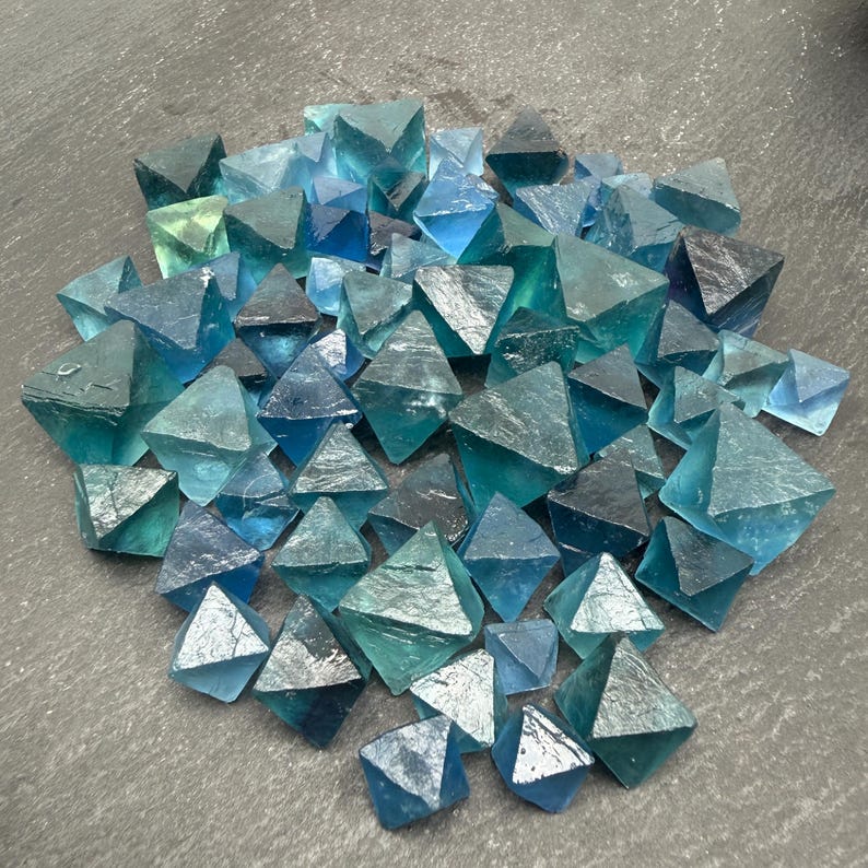 May include: A collection of numerous small, translucent blue and teal fluorite crystals, each shaped as an octahedron with sharp geometric edges and smooth, glassy surfaces, clustered together to display varying shades of deep ocean blue and light green.