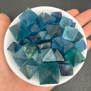 May include: A collection of small, translucent blue and teal fluorite crystals shaped into octahedrons. These geometric mineral specimens feature varying shades of deep ocean blue, light turquoise, and dark navy, all contained within a white bowl.