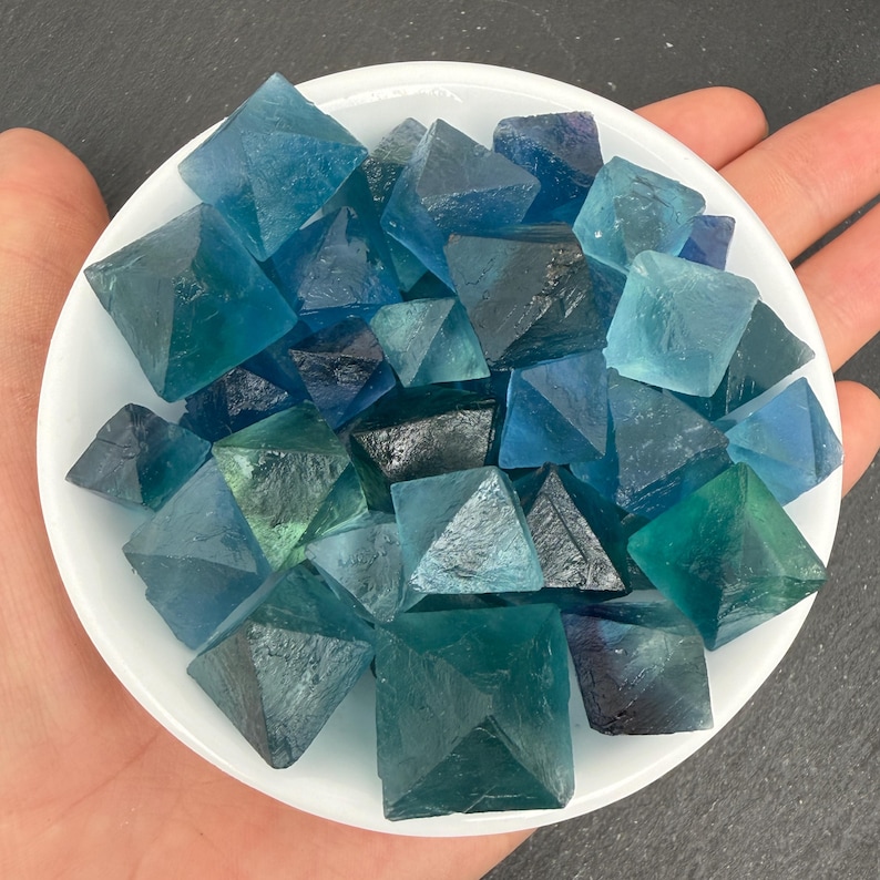 May include: A collection of small, translucent blue and teal fluorite crystals shaped into octahedrons. These geometric mineral specimens feature varying shades of deep ocean blue, light turquoise, and dark navy, all contained within a white bowl.