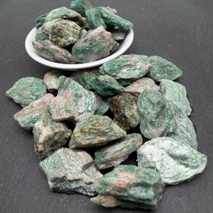 May include: A collection of raw green and pinkish-grey mineral stones, with some pieces contained in a small white bowl and others scattered around it. The stones have a rough, layered texture with visible crystalline surfaces and earthy tones.
