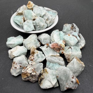 May include: A collection of raw, pale blue-green mineral stones with irregular shapes and textures. Some stones are contained within a small white bowl, while others are scattered around the base, displaying inclusions of brown and white mineral deposits.