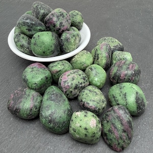 Peut inclure: Une collection de pierres de rubis sur zoïsite polies, présentant un mélange d'inclusions minérales vert vif et rouge magenta profond avec des taches noires. Certaines pierres sont contenues dans un petit bol blanc, tandis que d'autres sont éparpillées.