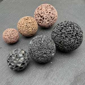 Pode incluir: Uma coleção de seis pedras vulcânicas esféricas de tamanhos variados. Três pedras são de cor castanho-claro ou castanho-avermelhado com uma textura porosa e cheia de orifícios, enquanto as outras três são cinzento-escuro ou pretas com uma superfície rugosa.