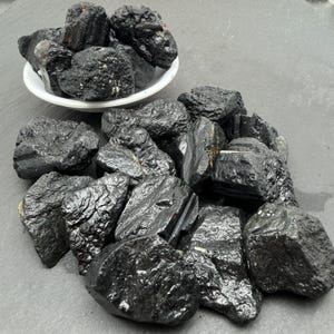 May include: A collection of raw black tourmaline stones with a glossy, crystalline texture and irregular, jagged shapes. Some stones are piled inside a small white bowl, while others are scattered in a cluster on a dark, flat surface.
