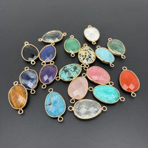 May include: A collection of teardrop-shaped gemstone charms, each encased in a gold-toned metal frame with two small loops at the top and bottom. The stones feature various colors and textures, including black, blue, green, pink, and white.