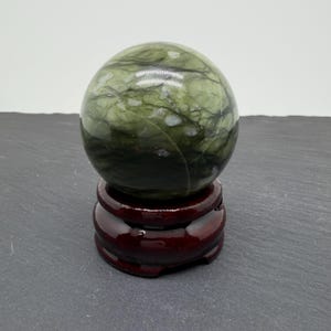 May include: A polished spherical stone in shades of sage and forest green with dark, intricate veining and white inclusions. The sphere rests securely on a circular, dark reddish-brown wooden display stand with a smooth, glossy finish.