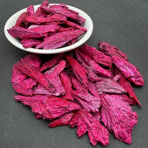 Op de afbeelding: Een verzameling fel magenta gekleurde kyaniet kristalsplinters, waarbij sommige stukken in een kleine witte kom zitten en andere eromheen verspreid liggen, met een duidelijke vezelige, mesachtige textuur en een metaalachtige glans.