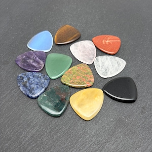 May include: A collection of twelve triangular guitar picks crafted from various polished stones, including blue, brown, red, purple, green, pink, white, black, yellow, and mottled mineral patterns, arranged in a cluster on a dark grey surface.