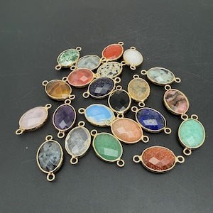 May include: A collection of oval-shaped jewelry connectors featuring faceted gemstones in various colors including green, orange, blue, black, and pink. Each stone is set in a gold-toned metal frame with two small loops on opposite ends.