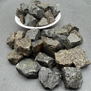 May include: A collection of rough, dark grey and charcoal-coloured granite stones with a speckled texture. Some stones are contained within a small white bowl, while others are scattered around the base, showcasing irregular shapes and jagged edges.