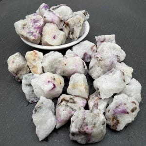 Puede incluir: Una colección de piedras de cuarzo blanco en bruto y sin pulir, con parches incrustados de minerales de color rosa vibrante y magenta. Algunas piedras están contenidas en un pequeño cuenco blanco, mientras que otras están esparcidas.