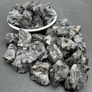 May include: A collection of raw, unpolished labradorite stones featuring a mottled grey and black colour palette with crystalline textures. Some stones are contained within a white ceramic bowl, while others are scattered around the base.