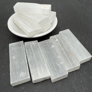 May include: A collection of rectangular selenite crystal bars with a translucent white appearance and fibrous texture. Several bars are stacked inside a small white ceramic bowl, while four additional bars are arranged in a row on the surface.