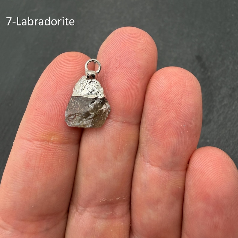 Pu&ograve; includere: Un ciondolo in Labradorite grezza con una pietra scura sfaccettata, una calotta metallica color argento e un piccolo anello circolare sulla parte superiore. Il testo 7-Labradorite &egrave; visibile nell'angolo in alto a sinistra.