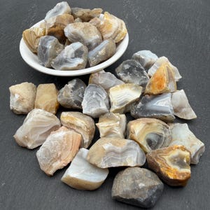 May include: A collection of raw, unpolished agate stones in shades of grey, white, tan, and amber. Many of the stones feature visible concentric banding patterns. Some stones are contained within a white bowl, while others are scattered nearby.