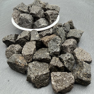 May include: A collection of dark gray, porous volcanic lava rocks with irregular shapes and rough textures. Some stones are contained within a small white circular dish, while others are scattered around the base of the dish on a flat surface.