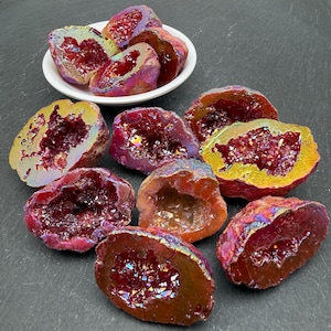 May include: A collection of deep red, iridescent geode halves with crystalline interiors. Several pieces are arranged in a small white bowl, while others are scattered around it, showcasing their rough, metallic-toned outer shells and sparkling centers.