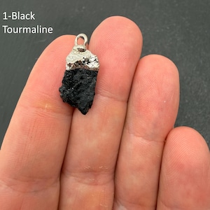 Pu&ograve; includere: Un ciondolo in tormalina nera grezza con una base in pietra scura, ruvida e testurizzata, sormontata da un gancio metallico color argento. Il testo 1-Black Tourmaline &egrave; visibile nell'angolo in alto a sinistra dell'immagine.
