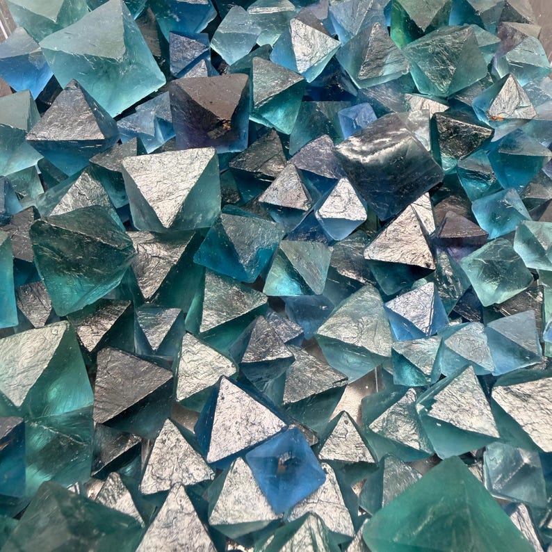 May include: A collection of numerous small, translucent blue fluorite crystals with a distinct octahedral shape. The crystals exhibit varying shades of teal, cyan, and deep navy blue, with some surfaces appearing smooth and others showing metallic luster.