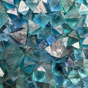 May include: A collection of numerous small, translucent blue and teal fluorite crystals. Each crystal features a distinct octahedral shape with sharp, geometric edges and smooth, glassy surfaces that reflect light, creating a dense, textured cluster.