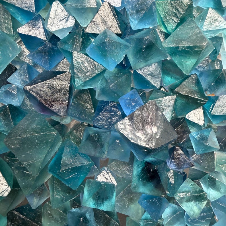May include: A collection of numerous small, translucent blue and teal fluorite crystals. Each crystal features a distinct octahedral shape with sharp, geometric edges and smooth, glassy surfaces that reflect light, creating a dense, textured cluster.