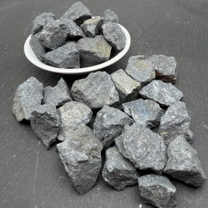 May include: A collection of raw, dark grey, metallic-looking mineral stones with jagged, uneven edges. Some stones are contained within a small, round, white ceramic bowl, while others are scattered around the base of the bowl.