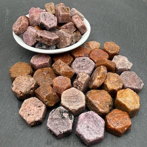 May include: A collection of raw, hexagonal-shaped corundum crystals in shades of pink, red, and brown. Some stones are contained within a small white bowl, while others are scattered around the base, displaying natural textures and earthy tones.