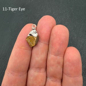 Pu&ograve; includere: Un piccolo ciondolo in pietra occhio di tigre grezza di colore marrone dorato con un gancio in metallo argentato sulla parte superiore. La pietra ha una consistenza naturale e ruvida, con l'etichetta 11-Tiger Eye nell'angolo in alto a sinistra.