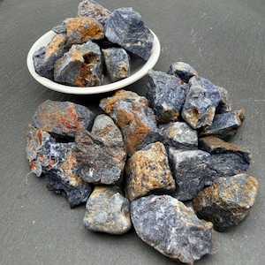 May include: A collection of raw, unpolished blue and brown stones, with some pieces contained in a small white bowl and others scattered around it. The stones feature irregular, jagged shapes with a mix of deep blue and earthy brown mineral tones.