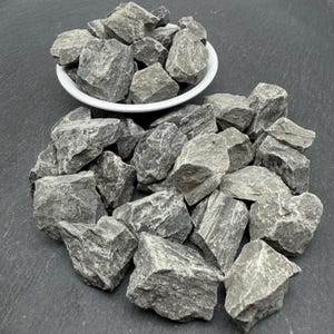 May include: A collection of rough, angular grey stones with varied textures and jagged edges. Some stones are contained within a simple white ceramic bowl, while others are scattered in a loose pile around the base of the bowl.