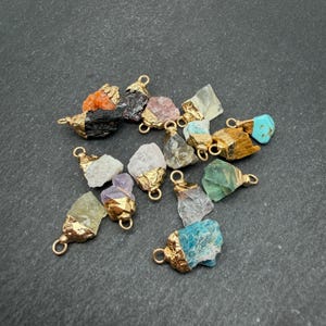 May include: A collection of raw, irregularly shaped gemstone pendants, each featuring a gold-toned metal cap with a small loop at the top. The stones display a variety of colors including orange, black, pink, white, brown, turquoise, and green.