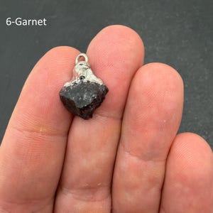 Pu&ograve; includere: Un ciondolo in granato grezzo rosso scuro con una calotta in metallo color argento e un piccolo occhiello circolare sulla parte superiore. La pietra ha una forma angolare irregolare con una superficie ruvida. Il testo 6-Garnet &egrave; visibile in alto a sinistra.