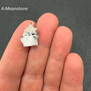 Pu&ograve; includere: Un ciondolo in pietra di luna grezza caratterizzato da una pietra bianca e grigia irregolare con inclusioni nere, sormontato da un gancio in metallo argentato. Il testo 4-Moonstone &egrave; visibile nell'angolo in alto a sinistra.