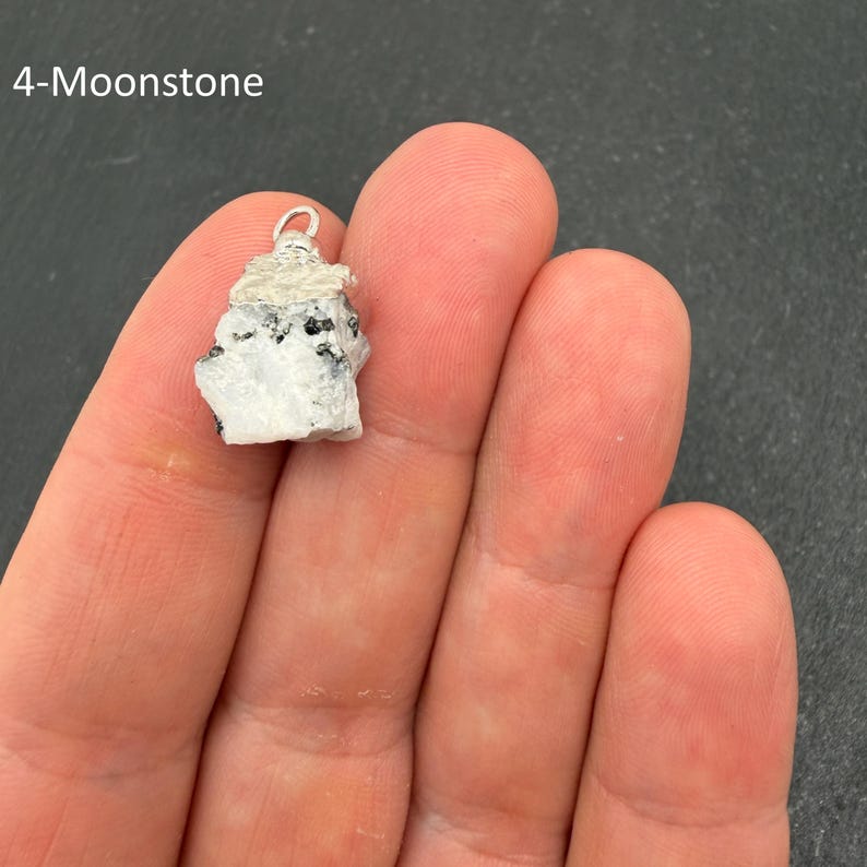 Pu&ograve; includere: Un ciondolo in pietra di luna grezza caratterizzato da una pietra bianca e grigia irregolare con inclusioni nere, sormontato da un gancio in metallo argentato. Il testo 4-Moonstone &egrave; visibile nell'angolo in alto a sinistra.
