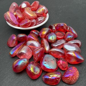 May include: A collection of polished, tumbled stones in shades of vibrant red and pink, featuring an iridescent, metallic sheen. Some stones are contained within a small white bowl, while others are scattered across the surface.