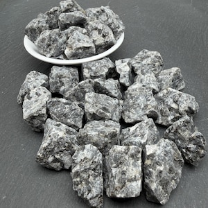 May include: A collection of raw, jagged grey and white stones with a marbled texture. Some stones are piled inside a small, round white ceramic bowl, while others are scattered loosely in front of the bowl, displaying natural, uneven edges.