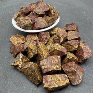 May include: A collection of raw, unpolished stones featuring a mottled pattern of deep red, mustard yellow, and earthy brown tones. Some of these jagged, angular mineral pieces are contained within a small white bowl, while others are scattered.