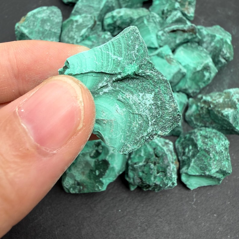 Op de afbeelding: Een verzameling ruwe malachietstenen met levendige groene tinten, opvallende bandpatronen en getextureerde oppervlakken. Elk stuk vertoont natuurlijke variaties in kleur en vorm, wat de karakteristieke gelaagde mineraalstructuur laat zien.