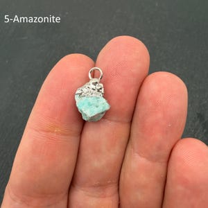 Pu&ograve; includere: Un ciondolo in pietra Amazzonite grezza di colore blu-verde chiaro con una calotta in metallo argentato testurizzato e un piccolo anello circolare sulla parte superiore. Il testo 5-Amazonite &egrave; visibile nell'angolo in alto a sinistra.