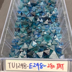 May include: A clear plastic rectangular container filled with numerous small, translucent blue and teal octahedral fluorite crystals. A white label on the front of the container displays the handwritten text TU1248-E248-280 TAT in black, blue, and red ink.